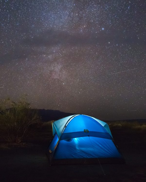 A lit dome tent on a hillside at night below a starry sky