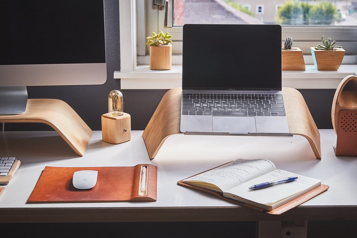 A tidy desk with a laptop on a wooden stand, a leather mouse pad and an open notebook