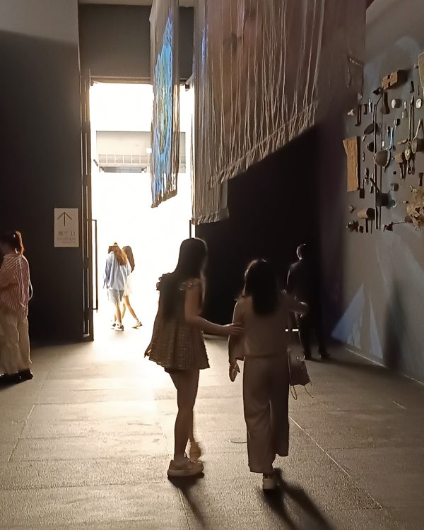 Visitors seen from behind walking down a dim gallery hall toward bright light