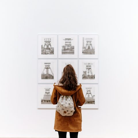 A visitor with a backpack seen from behind studying a grid of small prints on a white wall