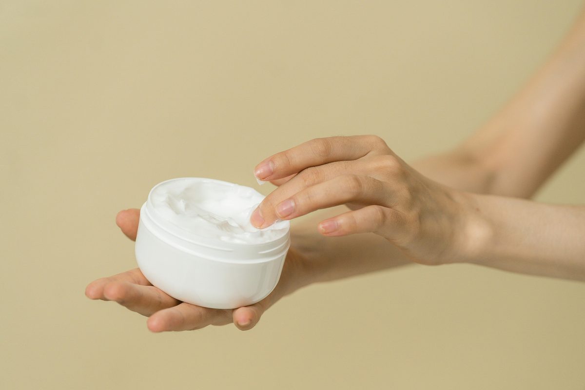 Two hands open a small white jar of face cream against a warm beige wall