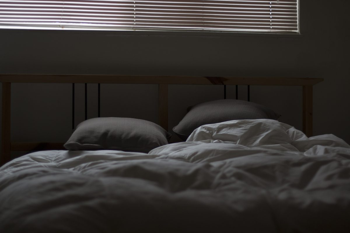 An unmade bed with grey sheets in a dim room with closed blinds