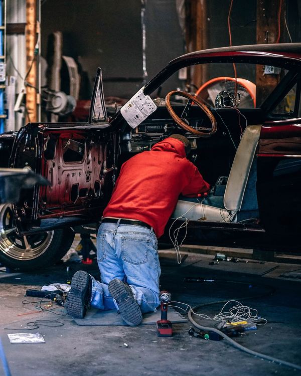 A mechanic in a red shirt kneels beside a car in a busy workshop