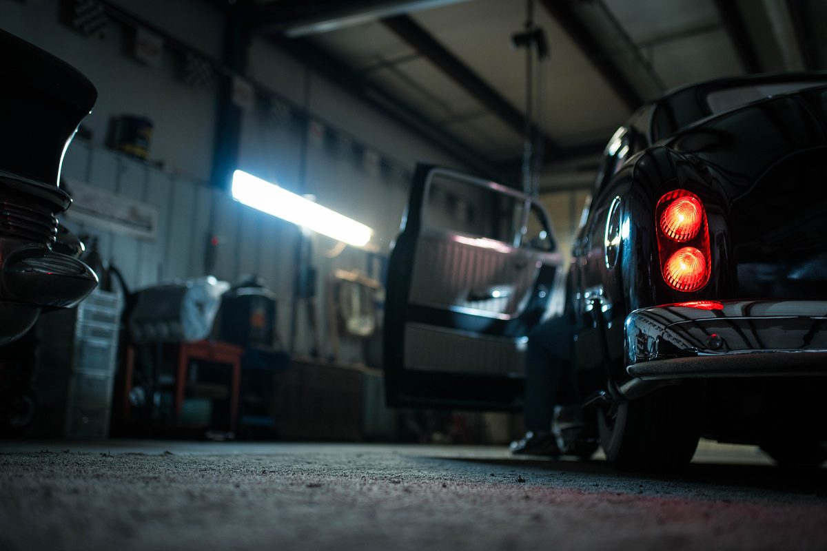 A dark classic car with red tail lights and an open door inside a quiet workshop