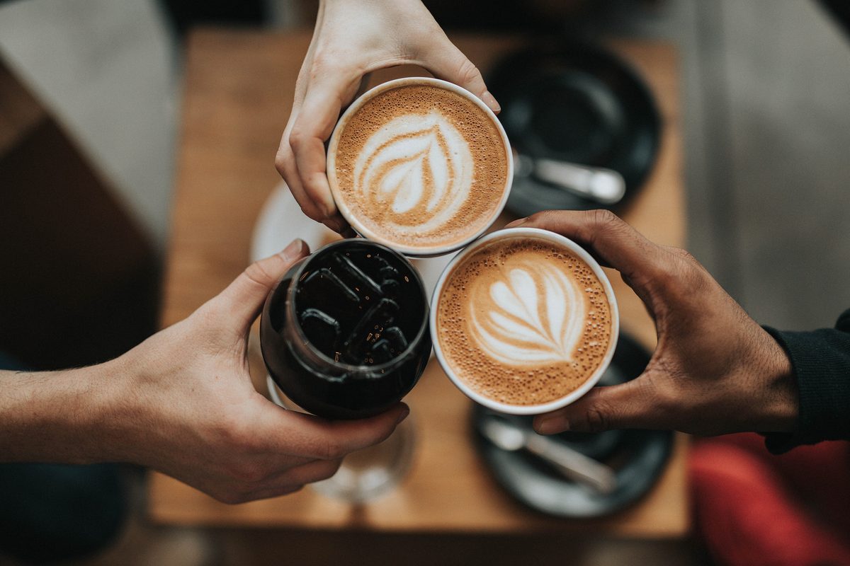 Three friends clink cups of latte with leaf art over a wooden cafe table