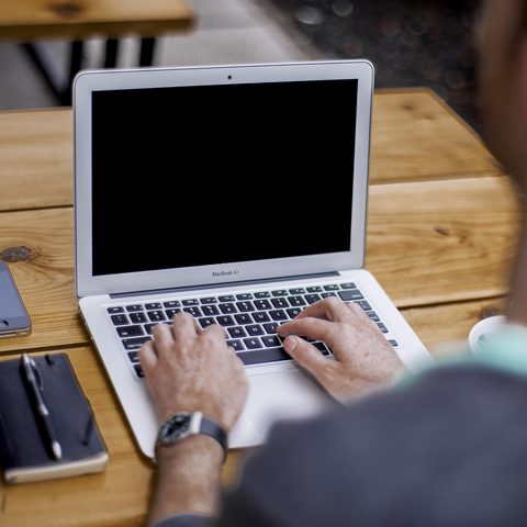 Hands typing on a laptop on a wooden desk, with a notebook beside it