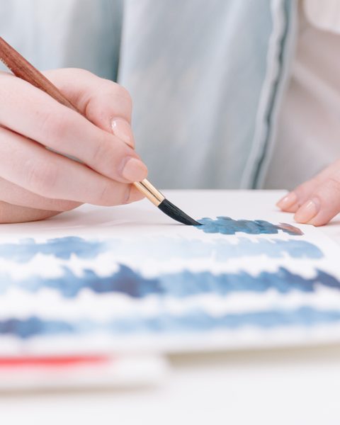 A hand paints soft blue watercolor strokes on white paper
