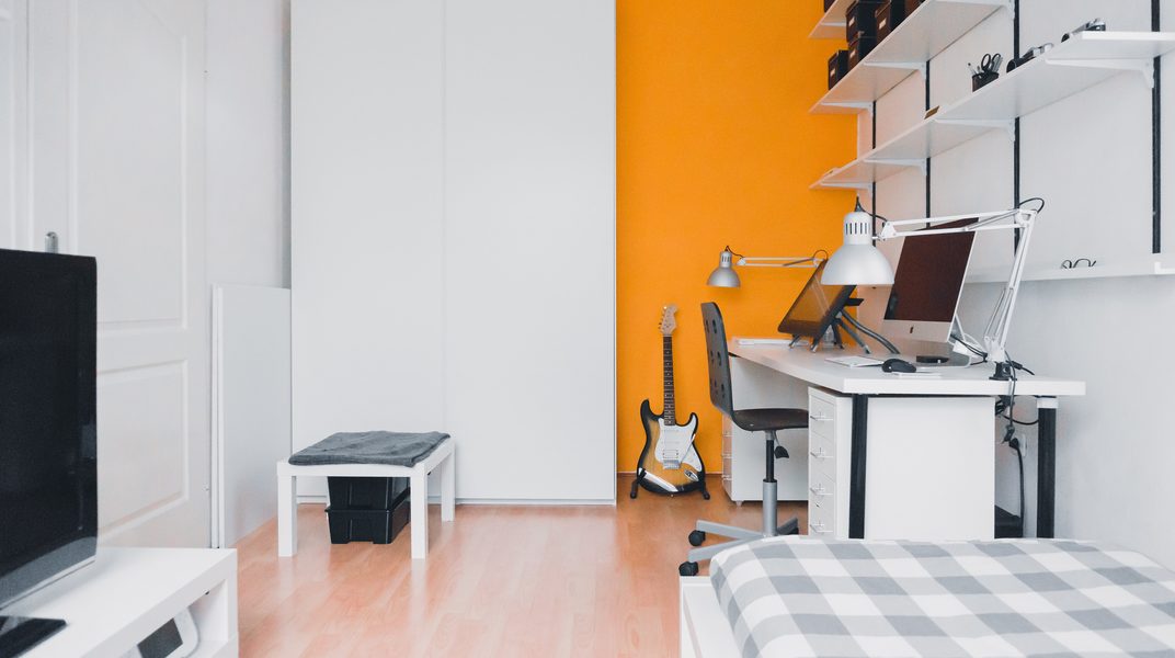 White room with an orange wall, a desk, a guitar and shelves