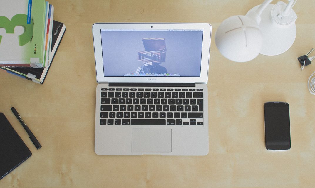 Laptop, notebook, pen and phone on a light wooden desk seen from above