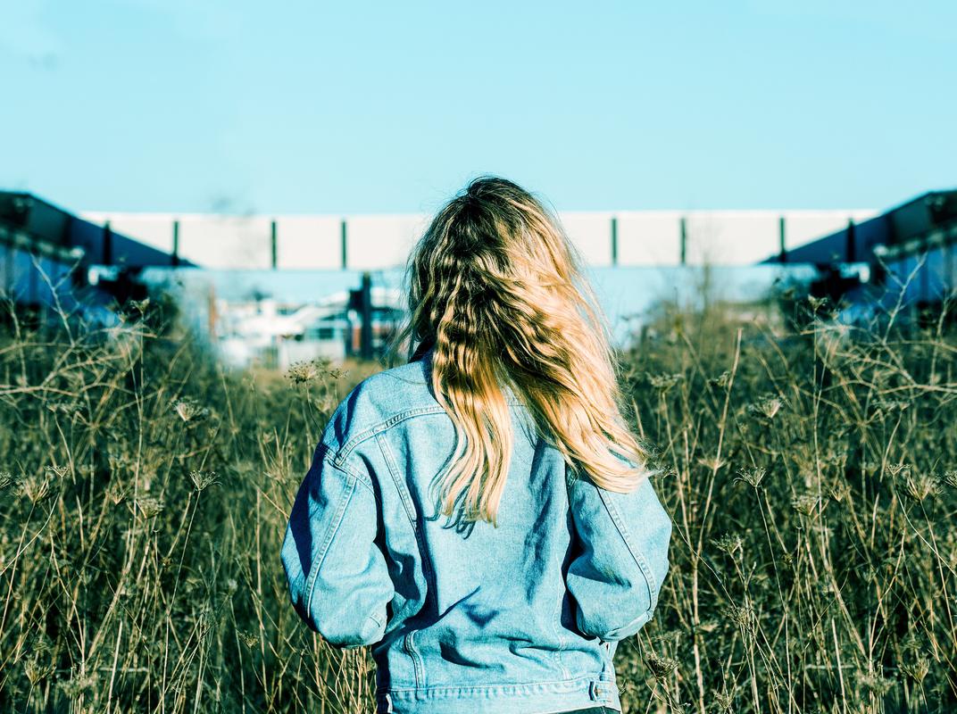 Woman in a light denim jacket walking away through tall grass under a blue sky