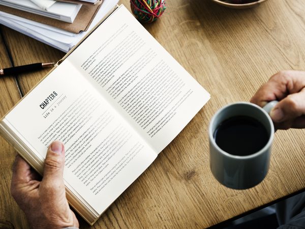 Hands hold an open paperback beside a mug of black coffee on a wooden table