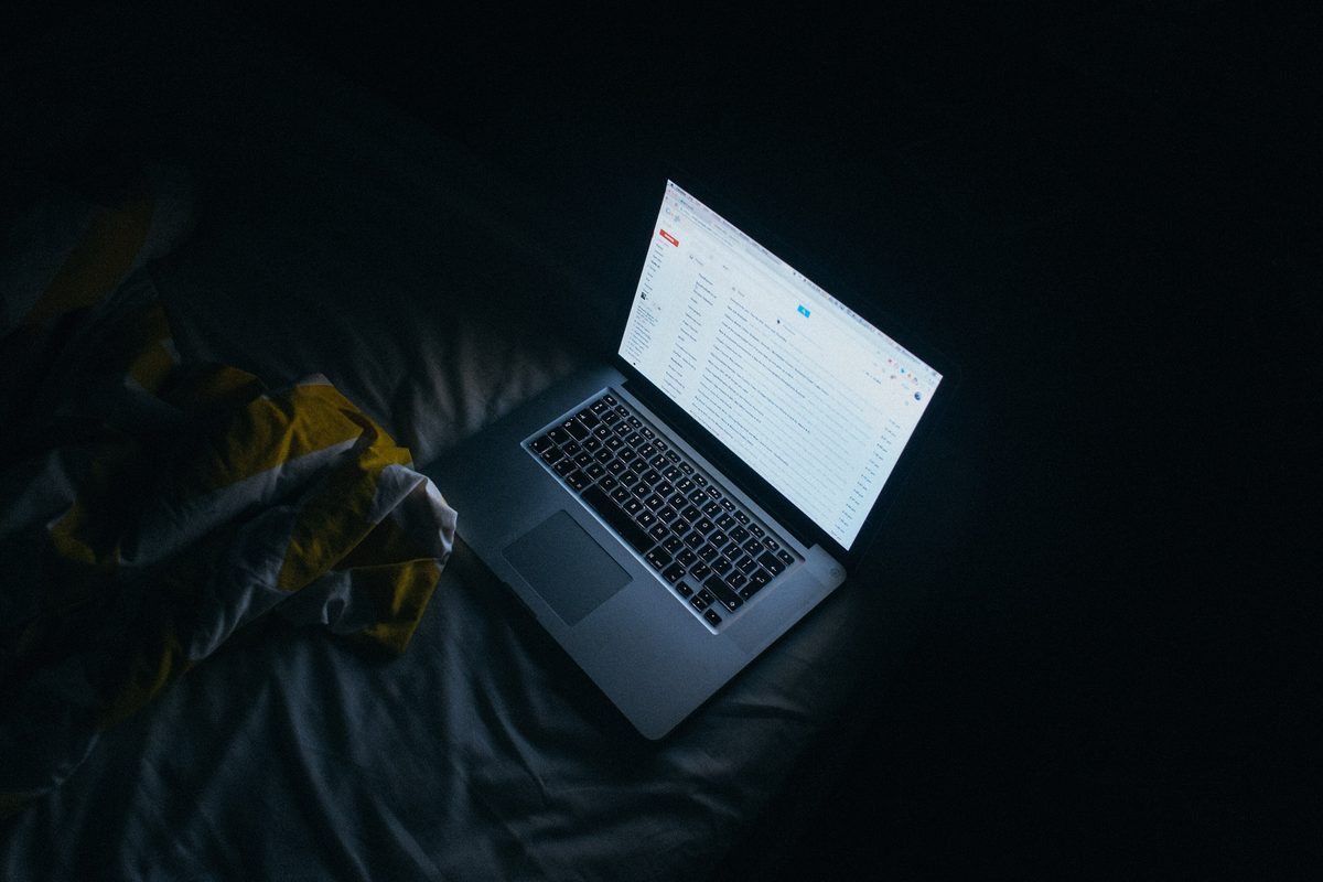 An open laptop glows on a dark bed in a room at night