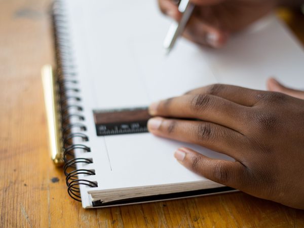 Hands with a pen and a ruler over an open spiral notebook on a wooden desk