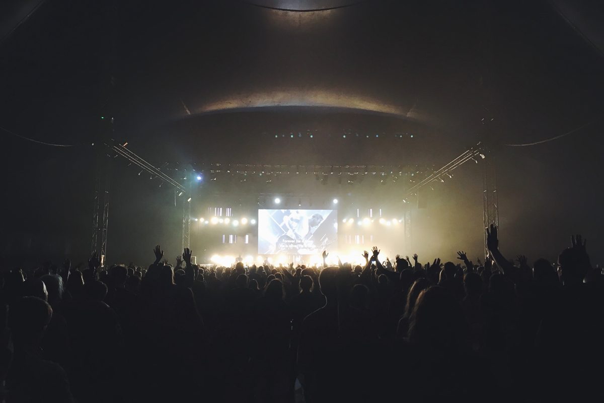 A crowd seen from behind in a dark hall facing a bright stage and screen