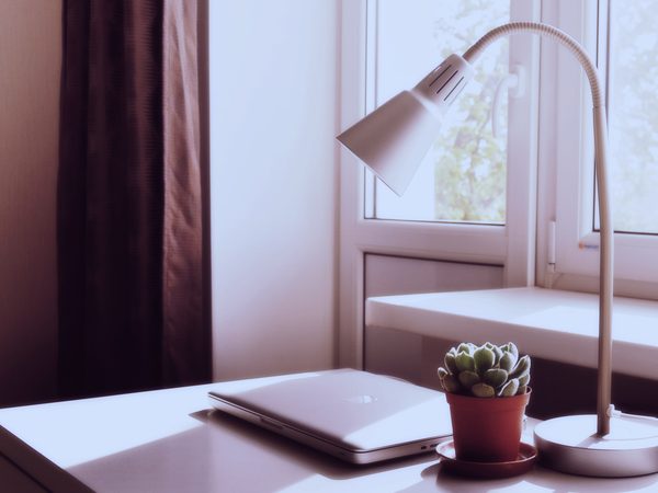 A closed laptop, a desk lamp and a small cactus by a bright window