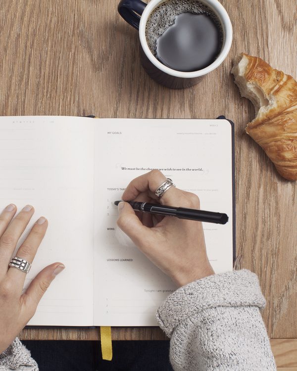 Hands writing in an open planner next to a cup of coffee and a croissant