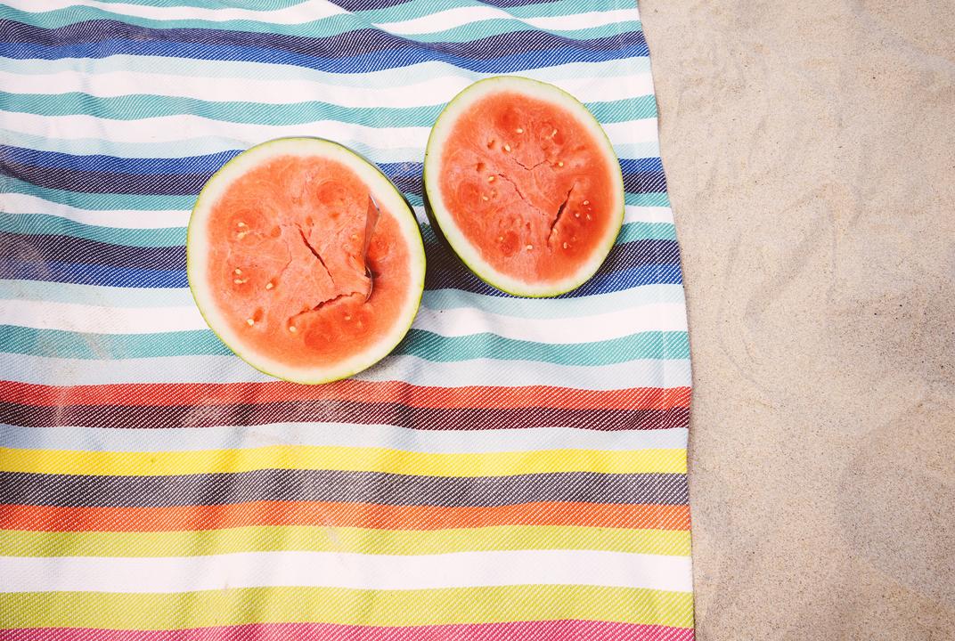 Two watermelon halves on a rainbow striped beach towel in the sand