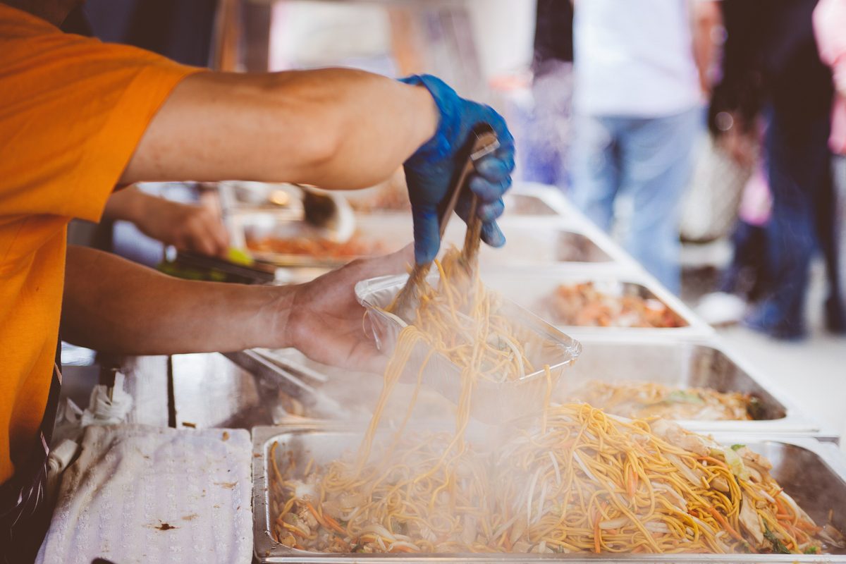 A volunteer in gloves serves steaming noodles from a long buffet tray