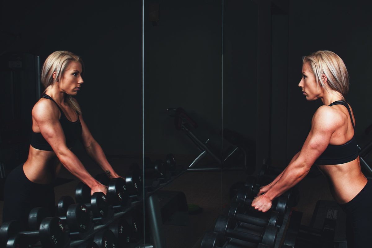 Two athletes reach for dumbbells on a rack in front of a dark gym mirror
