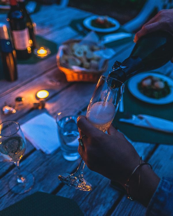Friends pouring sparkling wine at a candlelit outdoor table at dusk
