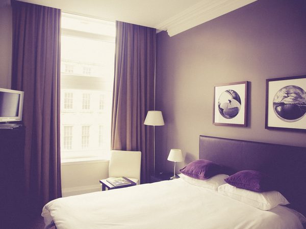 A quiet hotel bedroom with violet curtains, white bedding and a bright window