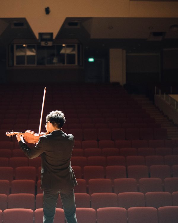 A musician seen from behind on stage facing rows of empty red seats