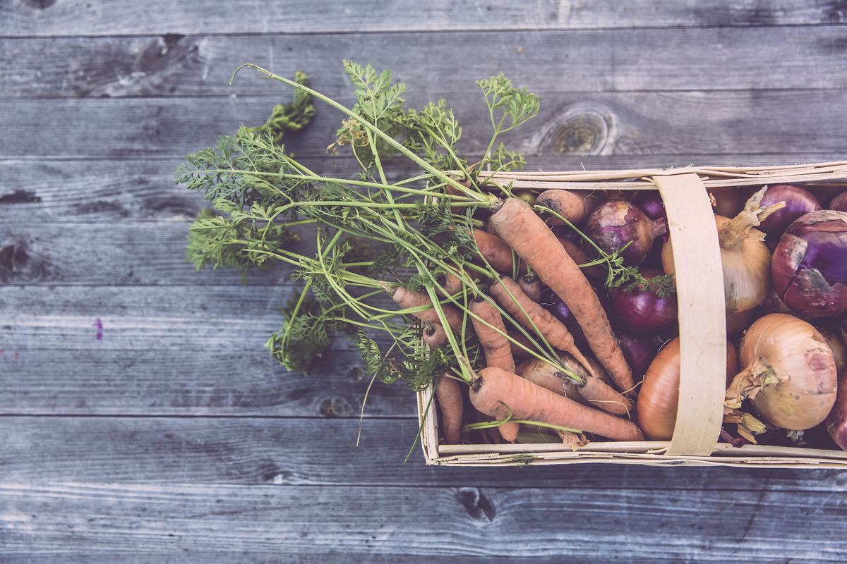 A wooden crate of carrots, beets and onions on grey boards seen from above