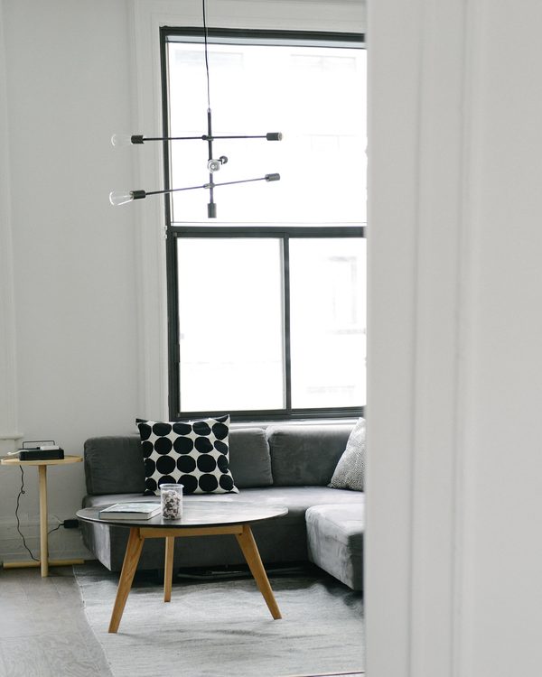 Grey corner sofa with a polka dot cushion and a round wood table by a tall window