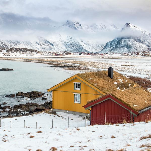 A yellow and a red house on a snowy shore below white mountains