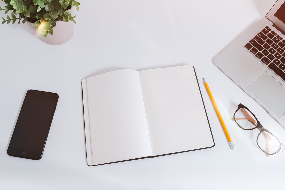 An open notebook, glasses, a pencil and a phone on a bright white desk