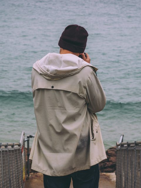 A person in a beige raincoat seen from behind at a pier railing over grey sea