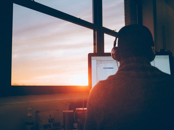 A listener in headphones seen from behind at a window at sunset