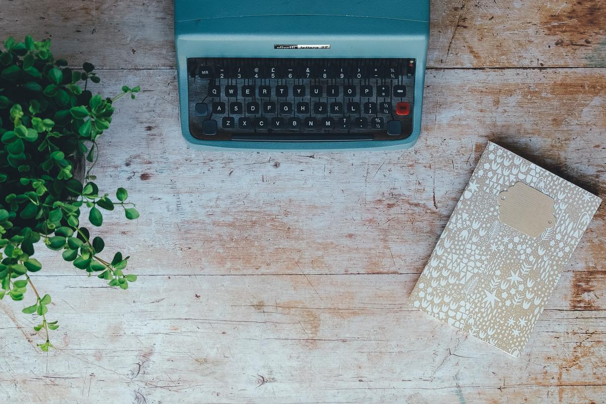 A teal typewriter, a patterned notebook and a plant on weathered wood seen from above