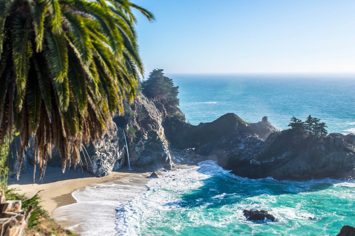 A turquoise cove below rocky cliffs, framed by palm leaves