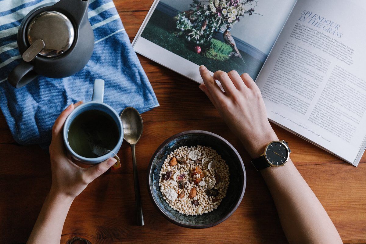 Hands hold a mug over a bowl of granola and an open magazine on a wooden table