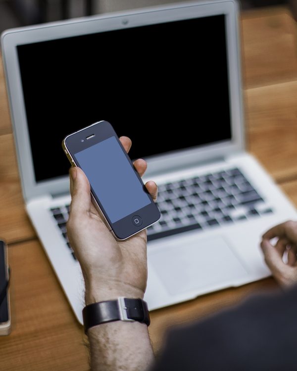 Hands holding a phone over an open laptop on a wooden desk