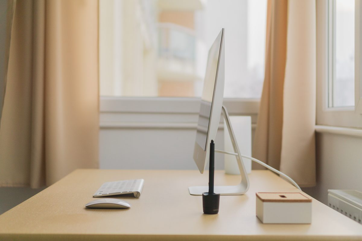 A slim desktop screen on a light wooden desk by a curtained window