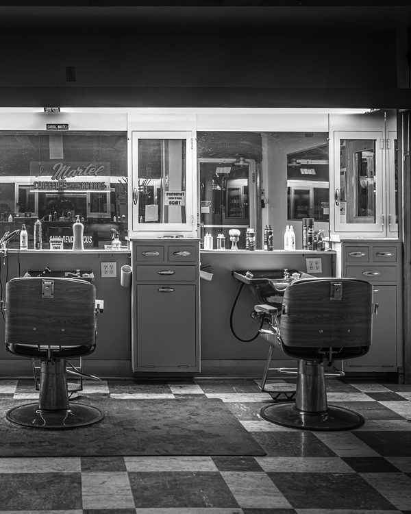 Two empty salon chairs in front of a wall of mirrors, in black and white