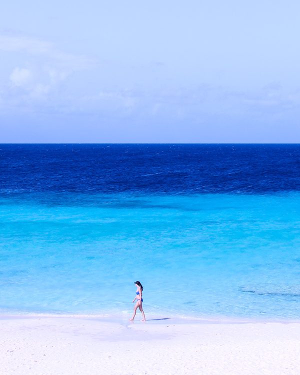 A lone swimmer walks along white sand beside bright turquoise water