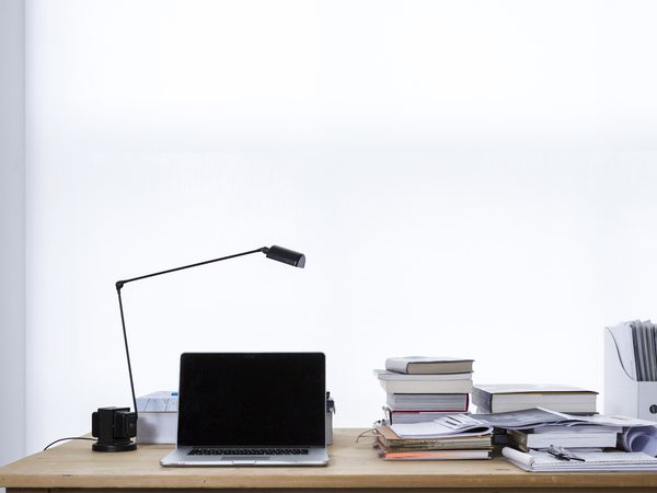 Laptop on a desk next to stacks of books and a lamp