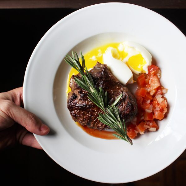 A hand holding a plate of braised beef with rosemary and roasted tomatoes