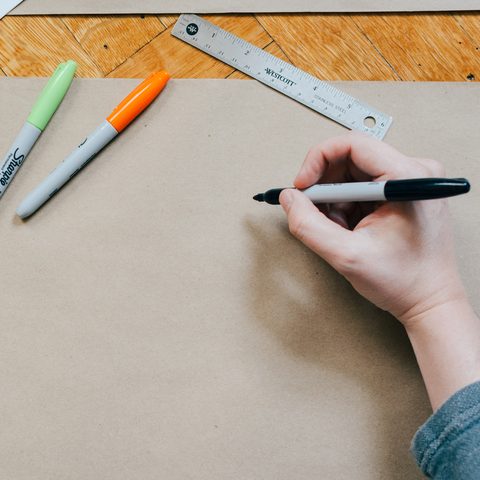 A hand draws with a black marker on brown paper next to a ruler