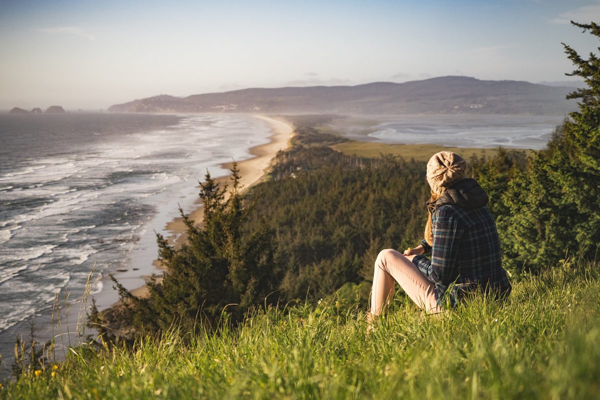 Woman in a plaid coat sitting on a grassy slope above a long sandy shore