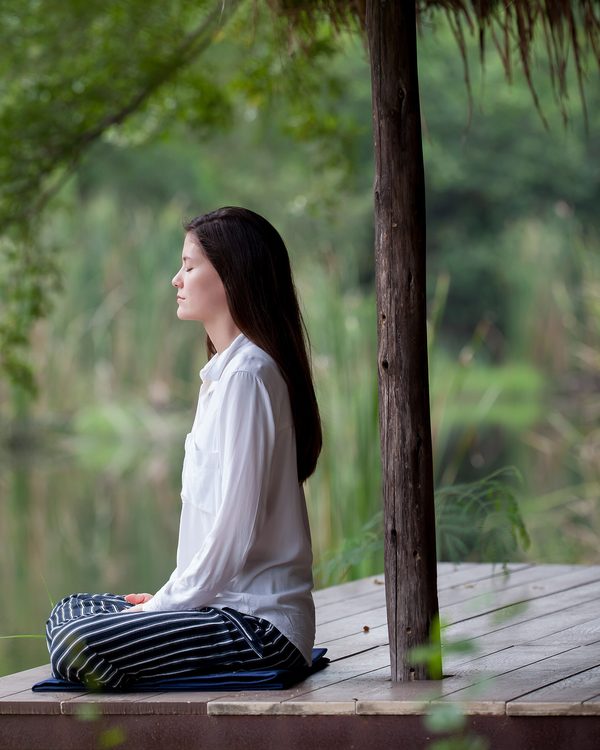 A woman sits cross legged on a wooden deck meditating beside a green lake