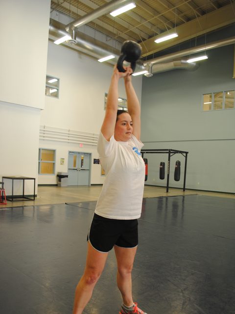 A woman pressing a kettlebell overhead in a bright gym
