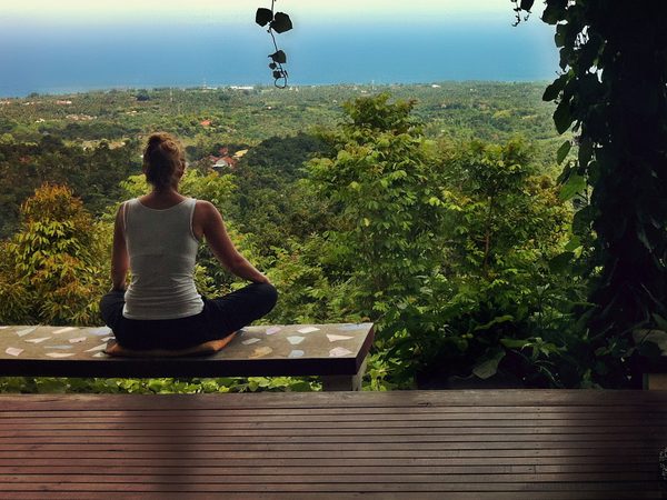 A woman meditates cross-legged on a bench above a green valley and the sea
